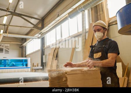 Giovane falegname in maschera protettiva mettendo sottili assi di legno in pila durante il suo lavoro alla fabbrica di mobili Foto Stock