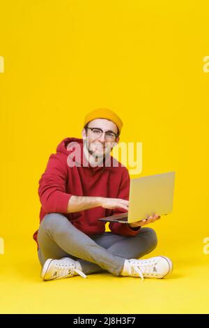 Happy giovane uomo hipster in rosso felpa con cappuccio seduta sul pavimento tenendo un computer portatile a studiare o a lavorare. Uomo caucasico freelance intelligente con notebook isolato su sfondo giallo. Foto Stock