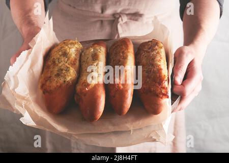 Mani di uomo che tengono caldo panino cotto su pane baguette con prosciutto, pancetta, verdure e formaggio su pergamena. Maschio in grembiule nero in casa cucina bac Foto Stock