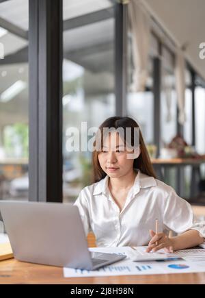 Una donna d'affari asiatica che lavora nella contabilità finanziaria analizza un grafico usando un calcolatore del laptop con i documenti e tenendo una penna seduta su una sedia a. Foto Stock