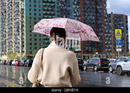 Pioggia in una città, donna in piedi con ombrello sulla strada della città sullo sfondo di edifici residenziali e auto parcheggiate Foto Stock