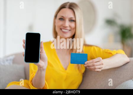 Felice bella giovane bionda caucasica donna in abiti gialli mostra smartphone con schermo vuoto e carta di credito Foto Stock