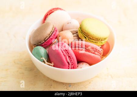 Macaroon multicolore e uova di cioccolato in ciotola di ceramica su sfondo di cemento beige. Vista laterale, primo piano, vita morta. Colazione, mattina, concetto. Foto Stock