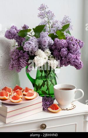 Un fragrante bouquet di lilacs colorati si erge in un vaso di vetro verde su sfondo bianco. Accanto a una tazza di tè caldo e affettato ​​grapefruit. Foto Stock