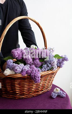 Giovane bel ragazzo mette in un cesto di legno un enorme bouquet colorato di lillacs. È in piedi su un tavolo coperto da una tovaglia viola Foto Stock