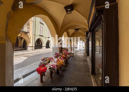 Busca, Cuneo, Italia - 13 maggio 2022: Antichi portici storici di via Umberto i con esposizione di fiori del negozio Foto Stock