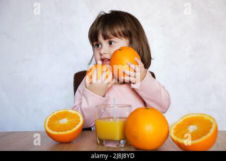 Ritratto di una bambina gioca con un arance su un tavolo. Ragazza piccola felice con un bicchiere di succo. Stile di vita sano. Foto Stock