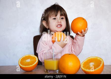 Ritratto di una bambina gioca con un arance su un tavolo. Ragazza piccola felice con un bicchiere di succo Foto Stock