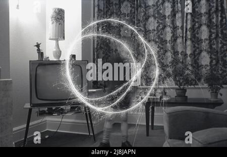 1970s, storico, un giovane schoooboy che gioca al chiuso in una sala con un scintillio di fuochi d'artificio, creando cerchi di luce bruciante, Inghilterra, Regno Unito. Fuochi d'artificio a mano, gli sparklers sono fatti da un filo metallico con un'estremità coperta in un combustibile di miscela di metallo, che quando acceso produce scintille brucianti lente. Tradizionalmente giocate con i bambini, sotto la supervisione di un adulto, prima di una festa o esposizione più grande dei fuochi d'artificio, gli scintille risalgono all'antica Grecia, dove erano originariamente creati come arma. Foto Stock