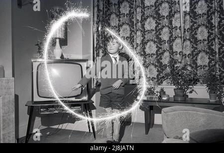 1970s, storico, un giovane schoooboy che gioca al chiuso in una sala con un scintillio di fuochi d'artificio, creando un cerchio di luce, Inghilterra, Regno Unito. Fuochi d'artificio a mano, gli sparklers sono fatti da un filo di metallo con un'estremità coperta in un combustibile di miscela di metallo, che quando illuminato producono scintille che bruciano lento. Tradizionalmente giocate con i bambini, sotto la supervisione di un adulto, prima di una festa o esposizione più grande dei fuochi d'artificio, gli scintille risalgono all'antica Grecia, dove erano originariamente creati come arma. Foto Stock