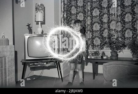 1970s, storico, un giovane schoooboy che gioca al chiuso in una sala con un scintillio di fuochi d'artificio, creando cerchi di luce, Inghilterra, Regno Unito. Fuochi d'artificio a mano, gli sparklers sono fatti da un filo di metallo con un'estremità coperta in un combustibile di miscela di metallo, che quando illuminato producono scintille che bruciano lento. Tradizionalmente giocate con i bambini, sotto la supervisione di un adulto, prima di una festa o esposizione più grande dei fuochi d'artificio, gli scintille risalgono all'antica Grecia, dove erano originariamente creati come arma. Foto Stock