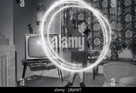 1970s, storico, un giovane schoooboy che gioca al chiuso in una sala con un scintillio di fuochi d'artificio, creando cerchi di luce bruciante, Inghilterra, Regno Unito. Fuochi d'artificio a mano, gli sparklers sono fatti da un filo metallico con un'estremità coperta in un combustibile di miscela di metallo, che quando acceso produce scintille brucianti lente. Tradizionalmente giocate con i bambini, sotto la supervisione di un adulto, prima di una festa o esposizione più grande dei fuochi d'artificio, gli scintille risalgono all'antica Grecia, dove erano originariamente creati come arma. Foto Stock