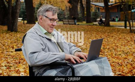 Felice anziano caucasico anziano maschio anziano nonno pensione siede da solo su sedia a rotelle maturo grigio-capelli pensionato solitario lavoro su laptop vincere online Foto Stock