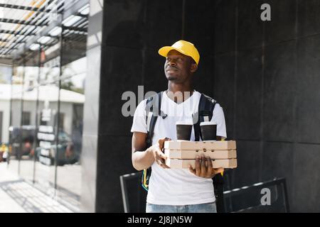 Uomo di consegna afro-americano in zaino giallo e tappo che contiene una pila di scatole di cartone per pizza con due tazze di caffè. Corriere maschio con pizza in attesa del destinatario. Foto Stock