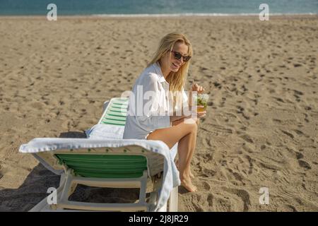 Giovane donna abbronzata in bikini e camicia con bevanda rinfrescante seduta su una sedia a sdraio, prendere il sole su una spiaggia Foto Stock