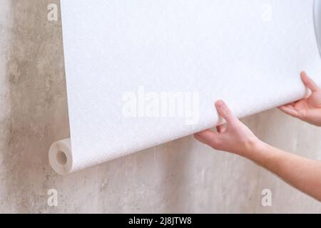 Un uomo tira fuori un rotolo di carta da parati bianca sulla parete con colla. Tappezzeria. Riparazione di una stanza, appartamento, casa. Foto Stock