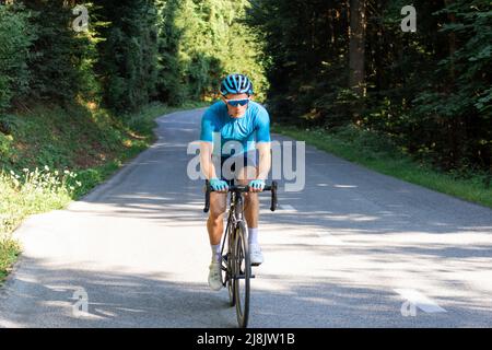 Ciclista maschile da corsa in una maglia blu sportiva, con casco e occhiali, che si sprint su una collina attraverso la foresta. Foto Stock