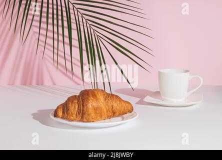 Foglia di palma tropicale, croissant fresco e una tazza di caffè su sfondo rosa pastello. Composizione minima degli alimenti. Concetto di colazione. Foto Stock