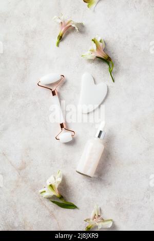 Rullo per il viso in giada bianca, gua sha, siero con fiori su tavola di pietra. Concetto di strumenti per la cura della pelle del viso. Foto Stock