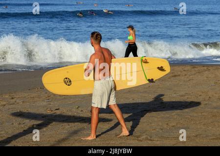 Un giovane uomo caucasico che trasporta una tavola da surf color crema alla spiaggia di Batu Bolong a Canggu, Bali, Indonesia, con una donna caucasica sullo sfondo. Foto Stock