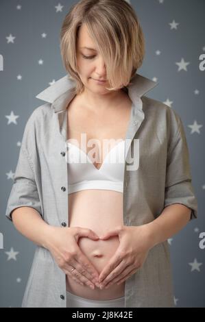 La donna incinta stringeva le mani a forma di cuore sul ventre. Bella femmina mostra cuore e segno d'amore sul suo bump bambino. Innamorato del nascituro. Foto Stock