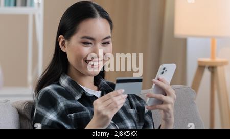 Sorridente etnia asiatica donna cliente utente in possesso di carta di credito bancario con denaro virtuale digitando smartphone a casa. Felice multietnico femmina shopper utilizzando Foto Stock