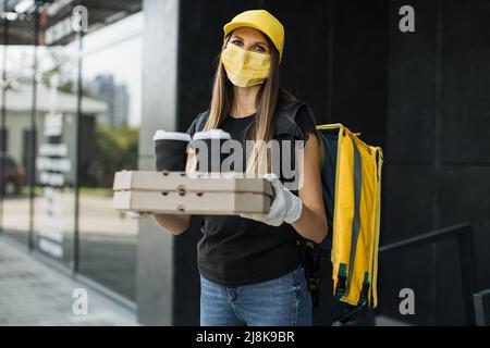 Ritratto di donna di consegna con cappuccio giallo in maschera facciale e zaino termico guardando la fotocamera. Corriere gioioso che consegna cibo veloce alla casa o all'ufficio del cliente. Consegna espressa. Foto Stock