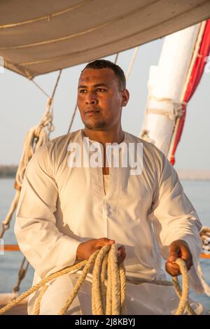 Membro dell'equipaggio che rifilatura le vele vestite con abiti tradizionali su una barca Nile Dahabiya Foto Stock