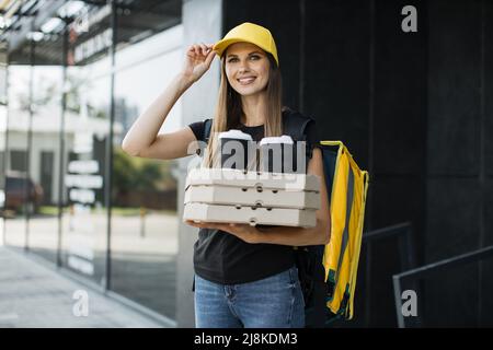 Donna di consegna caucasica con zaino giallo e tappo che contiene una pila di scatole di cartone per pizza con due tazze di caffè. Corriere femmina con pizza in attesa del destinatario. Foto Stock