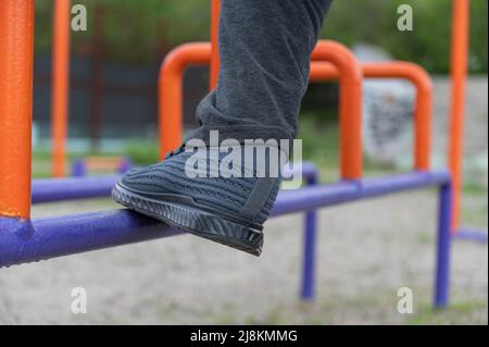 La parte inferiore di un uomo maturo impegnato in ginnastica rigenerativa sul terreno sportivo. Le gambe dell'uomo sono in piedi sulle barre blu e arancione. Reg Foto Stock