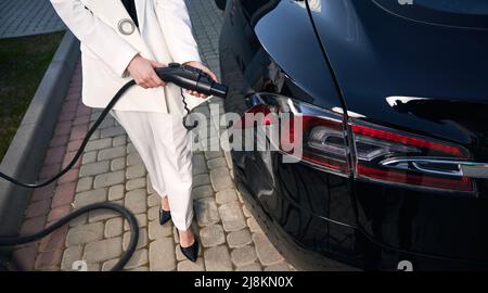 Primo piano di elegante giovane donna in tuta bianca carica auto elettrica su centrale elettrica. Donna d'affari che tiene il caricatore del veicolo elettrico mentre si trova in piedi accanto all'automobile sulla strada. Foto Stock