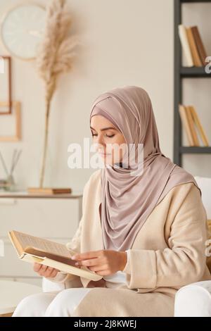 Bella giovane donna che indossa elegante hijab rosa pallido seduta in poltrona da solo in soggiorno lettura romanzo Foto Stock