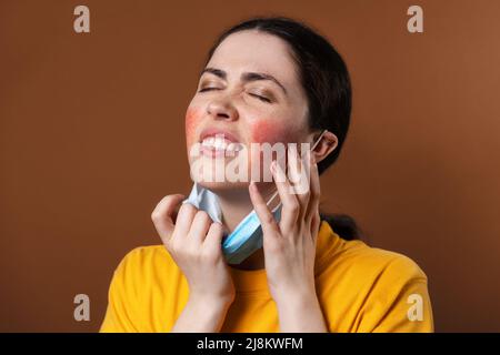 Rosacea. Una giovane donna turbata rigge nervosamente il viso con le mani per irritazione mentre indossa una maschera protettiva. Sfondo marrone. Il concep Foto Stock