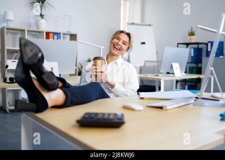 Donna di affari gioiosa con una tazza di caffè che mette le gambe sul tavolo in ufficio Foto Stock