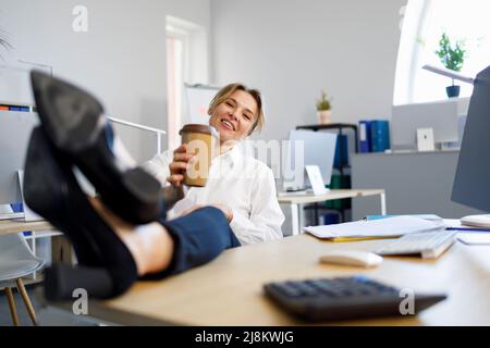 Donna d'affari che si rilassa durante la pausa caffè mettendo le gambe sulla scrivania Foto Stock