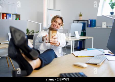 Una donna felice con una tazza di caffè rilassa mettendo le gambe sulla scrivania Foto Stock