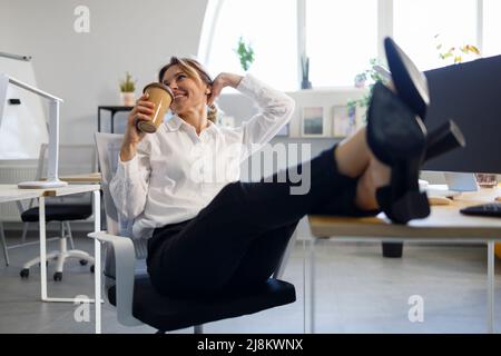 Felice donna d'affari che beve caffè mettendo le gambe sul tavolo Foto Stock