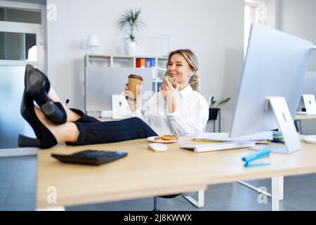 L'operaio dell'ufficio femminile beve caffè con biscotti per pranzo. Il boss femminile ha messo le gambe sulla scrivania Foto Stock
