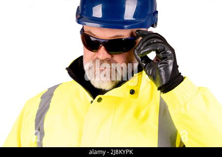 Un operaio in un cappello blu e un cappotto giallo hi-viz su sfondo bianco che indossa occhiali di sicurezza di colore scuro Foto Stock