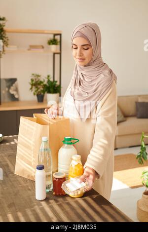 Verticale medio lungo colpo di bella giovane musulmana che indossa hijab in piedi a tavola che taking out cibo consegnato a lei Foto Stock