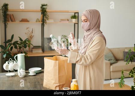 Donna musulmana che indossa hijab, maschera protettiva sul viso e guanti disinfettanti merci a casa dopo aver fatto shopping Foto Stock