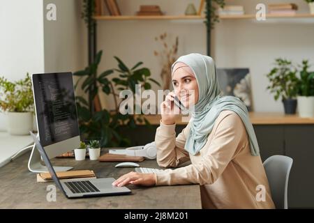 Ragazza allegra che indossa un hijab azzurro seduto al tavolo che parla al telefono mentre lavora al computer Foto Stock