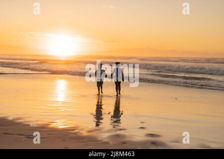 Vista posteriore dei fratelli afroamericani che tengono le mani e camminano sulla spiaggia contro il cielo al tramonto Foto Stock
