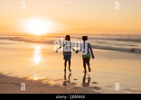 Vista posteriore dei fratelli afroamericani che tengono le mani e corrono sulla spiaggia contro il cielo al tramonto Foto Stock