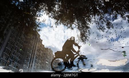 Silhouette sfocata di riflesso di una persona su una bicicletta da sola sul marciapiede bagnato della città in una giornata piovosa. L'aereo vola attraverso il cielo. Astratto p Foto Stock