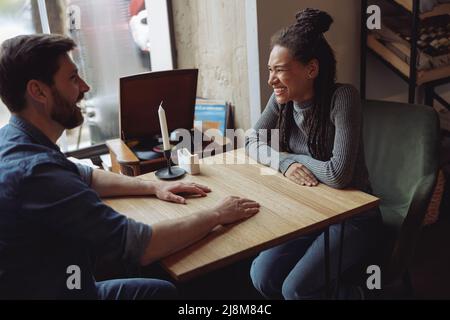 Allegro coppia di corse miste ridendo e chiacchierando al tavolo in un bar. Felice uomo e donna all'incontro. Foto Stock