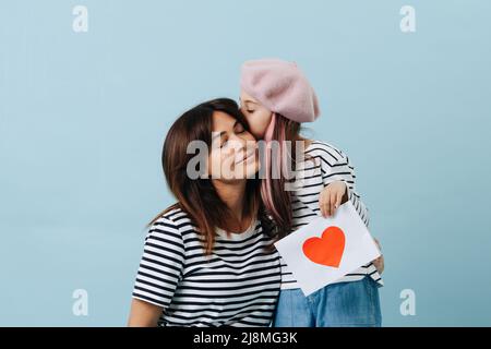 Tween ragazza in beretto francese baciando la mamma, lei sta tenendo un foglio di carta con cuore scarlatto su di esso. Su sfondo blu. Foto Stock