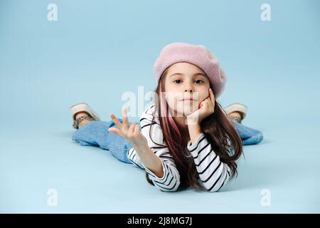 Ragazza cool moda Tween in beretto francese sdraiato sul pavimento, mostrando segno V. Ha tinto i capelli ciocche. Posa su sfondo blu. Foto Stock