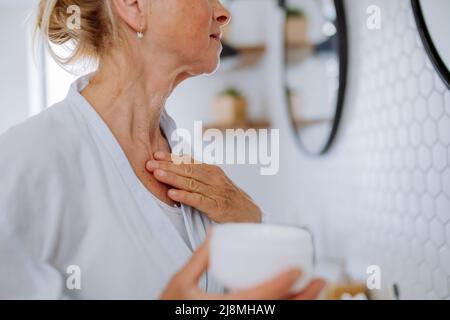 Bella donna anziana in accappatoio applicazione crema naturale in bagno, cura della pelle e mattina concetto di routine. Foto Stock