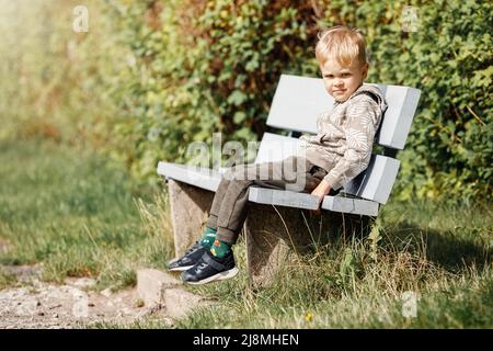 Un ragazzino sorridente si siede su una panca grigia in legno in un parco cittadino sullo sfondo del verde fogliame estivo e della luce del sole. Foto Stock